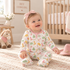 A baby girl sitting on a cream rug in a nursery, wearing a white footed sleeper with a pink collar and a colorful print of boots, hats, and trees, complete with a pink bow headband.