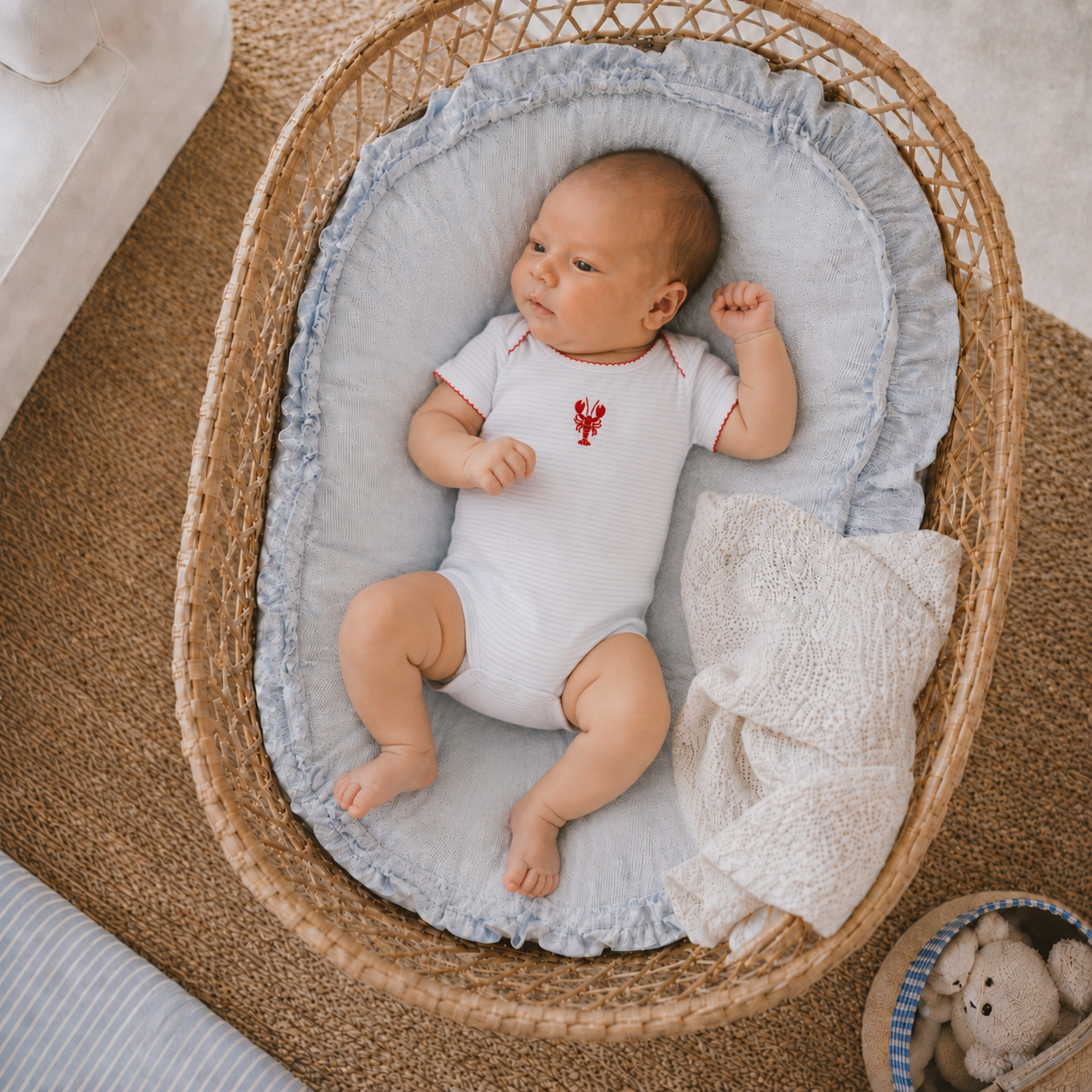 Baby in a white onesie with a red lobster hand embroidery design lying on a textured blanket in a wicker basket.