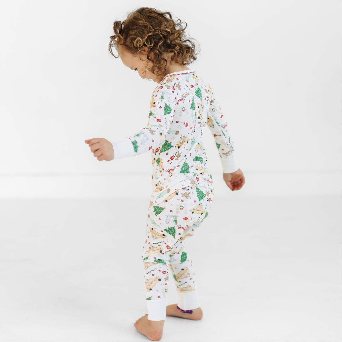 A toddler with curly brown hair wearing a white long-sleeve pajama set with a colorful Christmas print, standing barefoot against a plain white studio background.