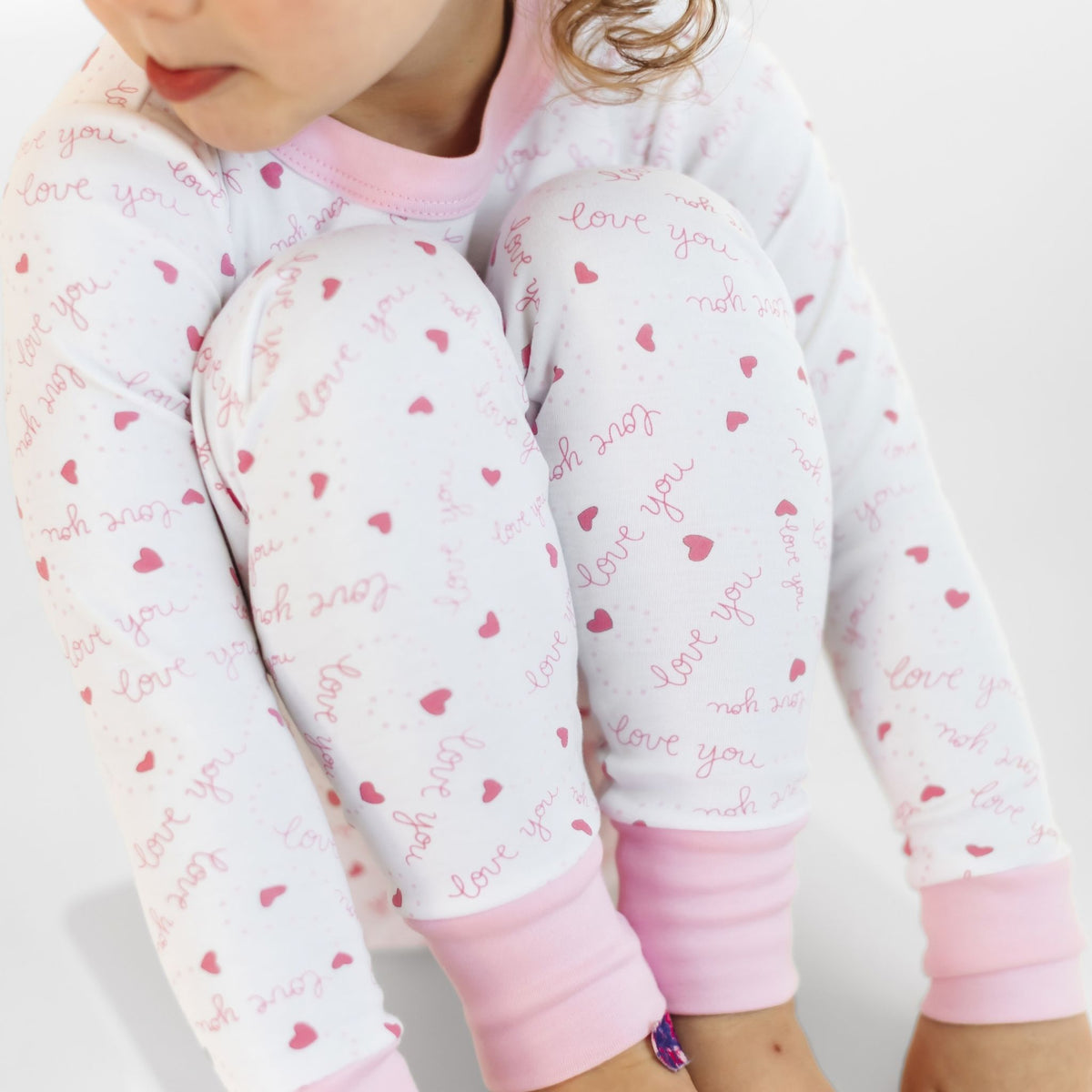 A child wearing a white long-sleeve pajama set with pink cuffs and a repeating pattern of pink hearts and cursive 'love you' text, shown in a close-up studio shot.
