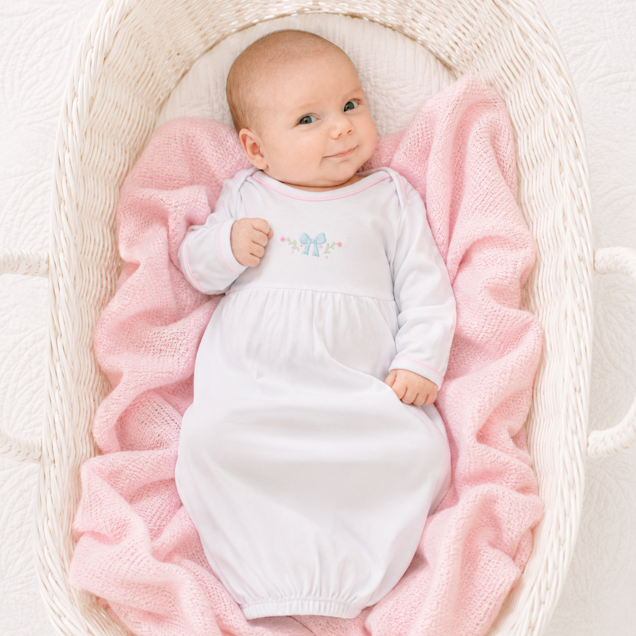 Baby in a white hand embrdoiered lgown by magnolia baby ying on pink blankets in a white bassinet