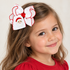 A young girl with long light brown hair wearing a large white hair bow with red trim and a Santa Claus face applique, paired with a red shirt against a plain light background.