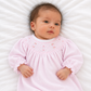 A baby lying on a white textured blanket, wearing a light pink long-sleeve baby smocked dress with tiny embroidered details on the chest.
