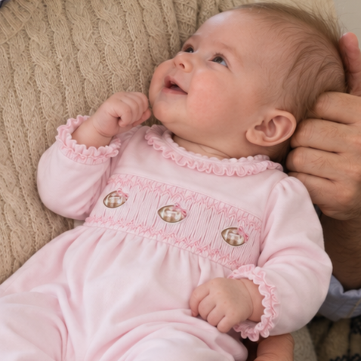 A baby girl wearing a light pink long-sleeve smocked jumpsuit with ruffled trim and three footballs on smocking lying on a textured beige knit blanket with her dads hand supporting the baby.