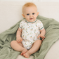 A baby sitting on a soft green muslin blanket, wearing a white short-sleeve bodysuit with a mallard duck print and green trim.