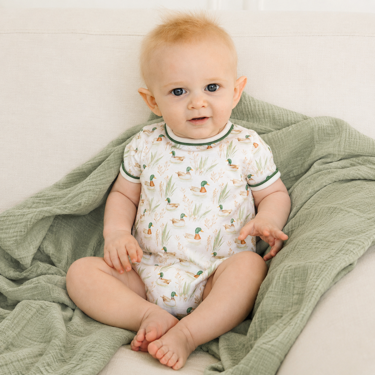A baby sitting on a soft green muslin blanket, wearing a white short-sleeve bodysuit with a mallard duck print and green trim.